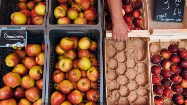 A market display of apples in crates and a rack of eggs and fruit, with a hand reaching for apples, and a chalkboard sign nearby, pretty and colorful.