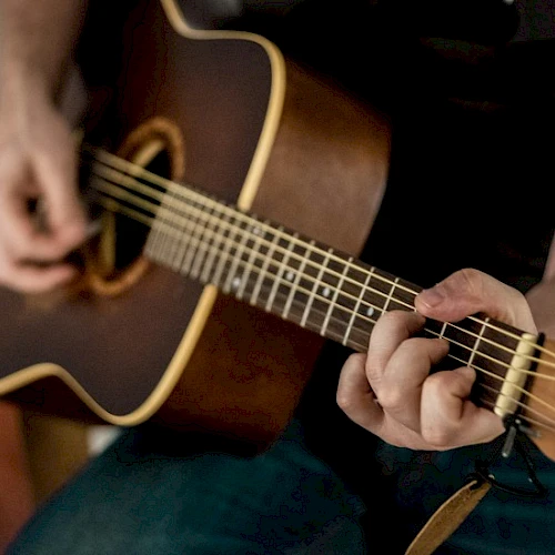 A person playing an acoustic guitar, fingers on the fretboard, strumming near the soundhole, close-up of hands and guitar neck.