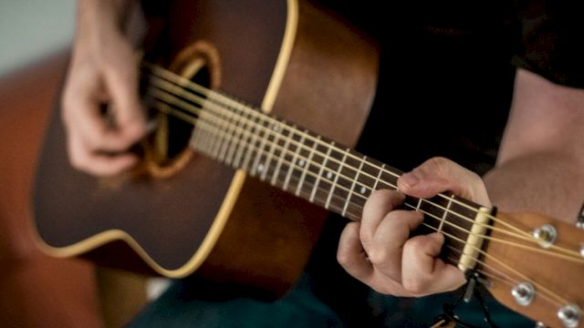 A person playing an acoustic guitar, fingers on the fretboard, strumming near the soundhole, close-up of hands and guitar neck.