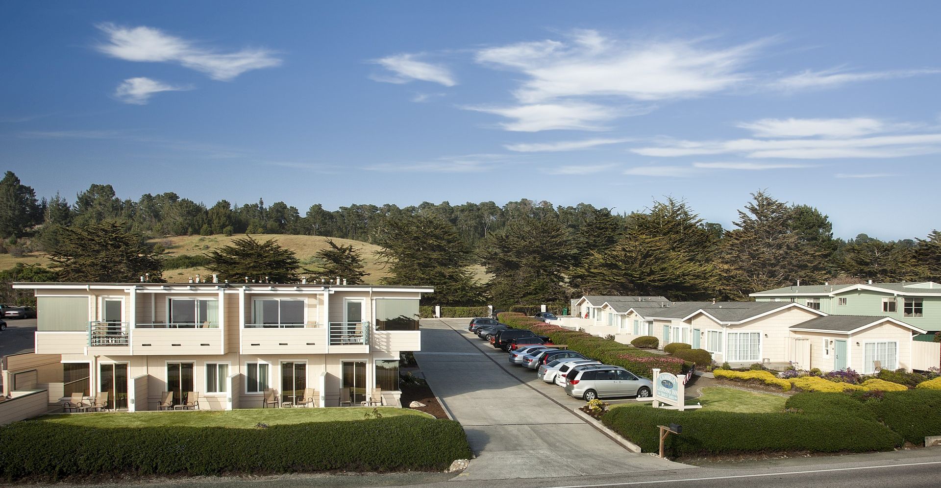 A row of modern two-story apartments with a shared parking lot, green hedges, and a backdrop of trees under a blue sky with light clouds.