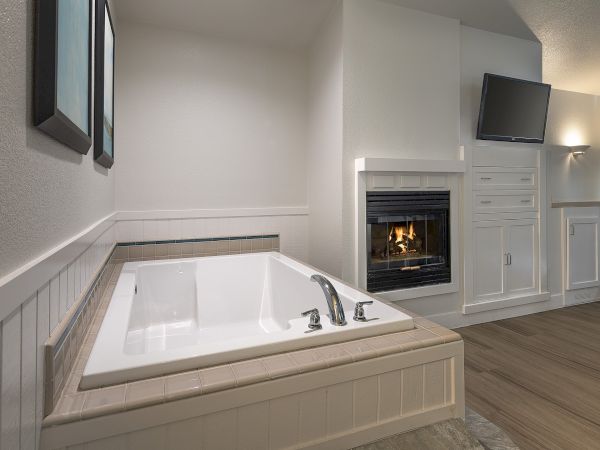 A modern bathroom with a built-in soaking tub, white paneling, a gas fireplace, and a wall-mounted TV above a white cabinet.