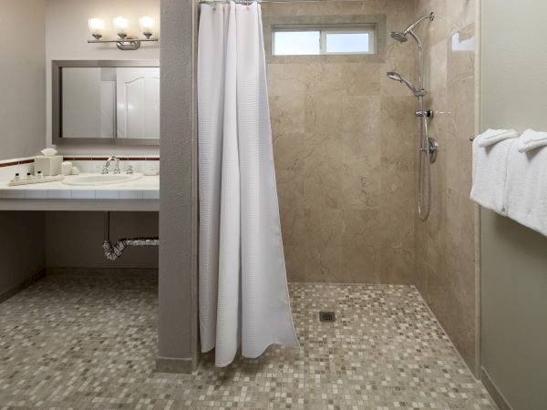 A spacious bathroom with a tiled walk-in shower, a white curtain, a double sink vanity, beige walls, and a towel rack on the right.