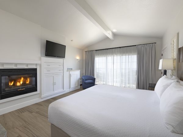 A bright hotel room with a lit fireplace, wall-mounted TV, white cabinetry, a large bed, blue chair, and a sliding glass door with curtains.