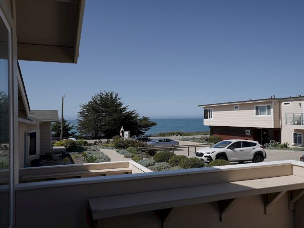 A sunny coastal view of a beach town: rooftops, parked cars, shrubs, and a clear blue ocean horizon beyond.