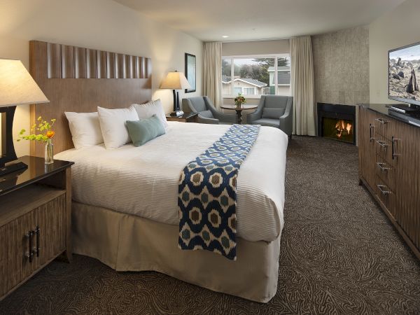 A cozy hotel room with a large bed, two nightstands, a seating area by the window, a fireplace, and a flat-screen TV on a dresser.