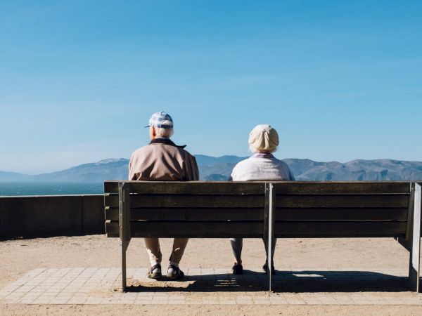Two elderly people sit on a bench facing the water, enjoying a peaceful seaside view under a clear blue sky.