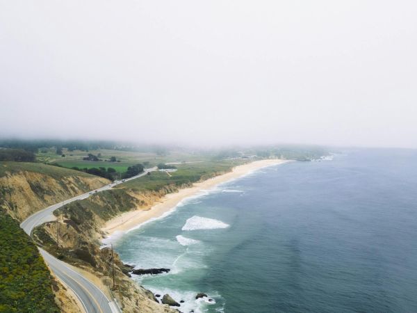 Coastline curves along with a winding road beside the ocean, waves grazing sandy beaches under a misty, low-lying horizon.