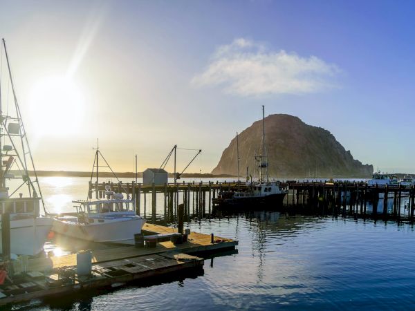 Harbor scene at sunset with boats docked by a wooden pier; a rocky island rises in the background, calm water reflecting the sky.