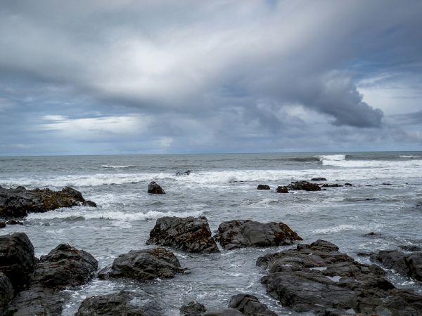 Rugged coastline with dark rocks, gray waves crashing, and a cloudy, moody sky over the sea.