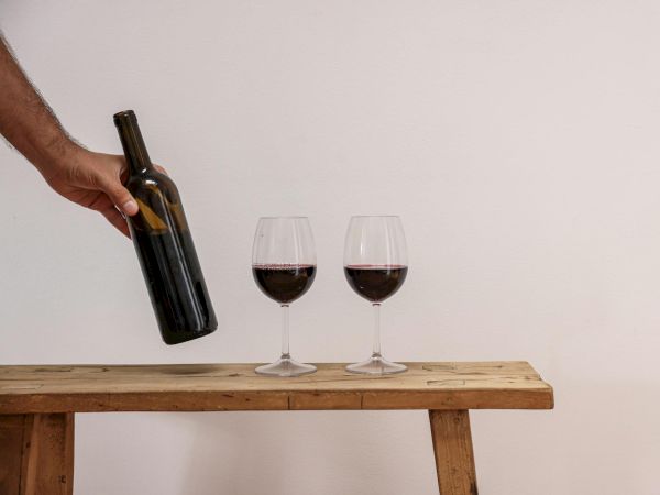 A hand pours red wine into two glasses on a wooden bench, ready for a simple, quiet tasting moment.