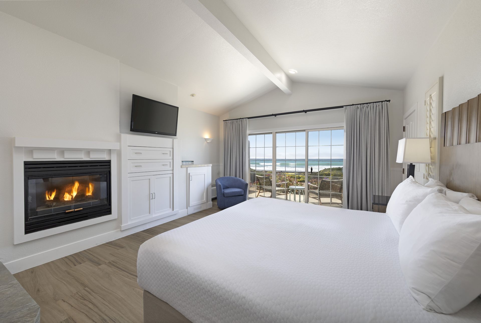 A bright hotel room with a fireplace, mounted TV, built-in cabinets, a large bed, blue chair, and a balcony with ocean view, ending with a period.