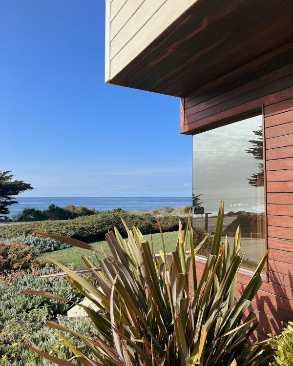 A modern house with brick siding and a large window overlooks a garden and the ocean in the distance, under a clear blue sky.