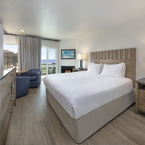 A modern hotel room with a queen bed, two nightstands, a flat-screen TV, a desk, a blue sofa, and a balcony with a seaside view, bright and airy.