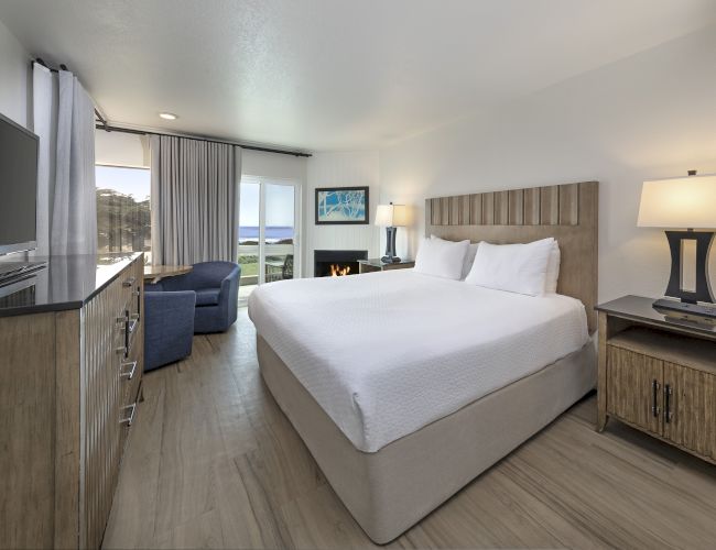 A modern hotel room with a queen bed, two nightstands, a flat-screen TV, a desk, a blue sofa, and a balcony with a seaside view, bright and airy.