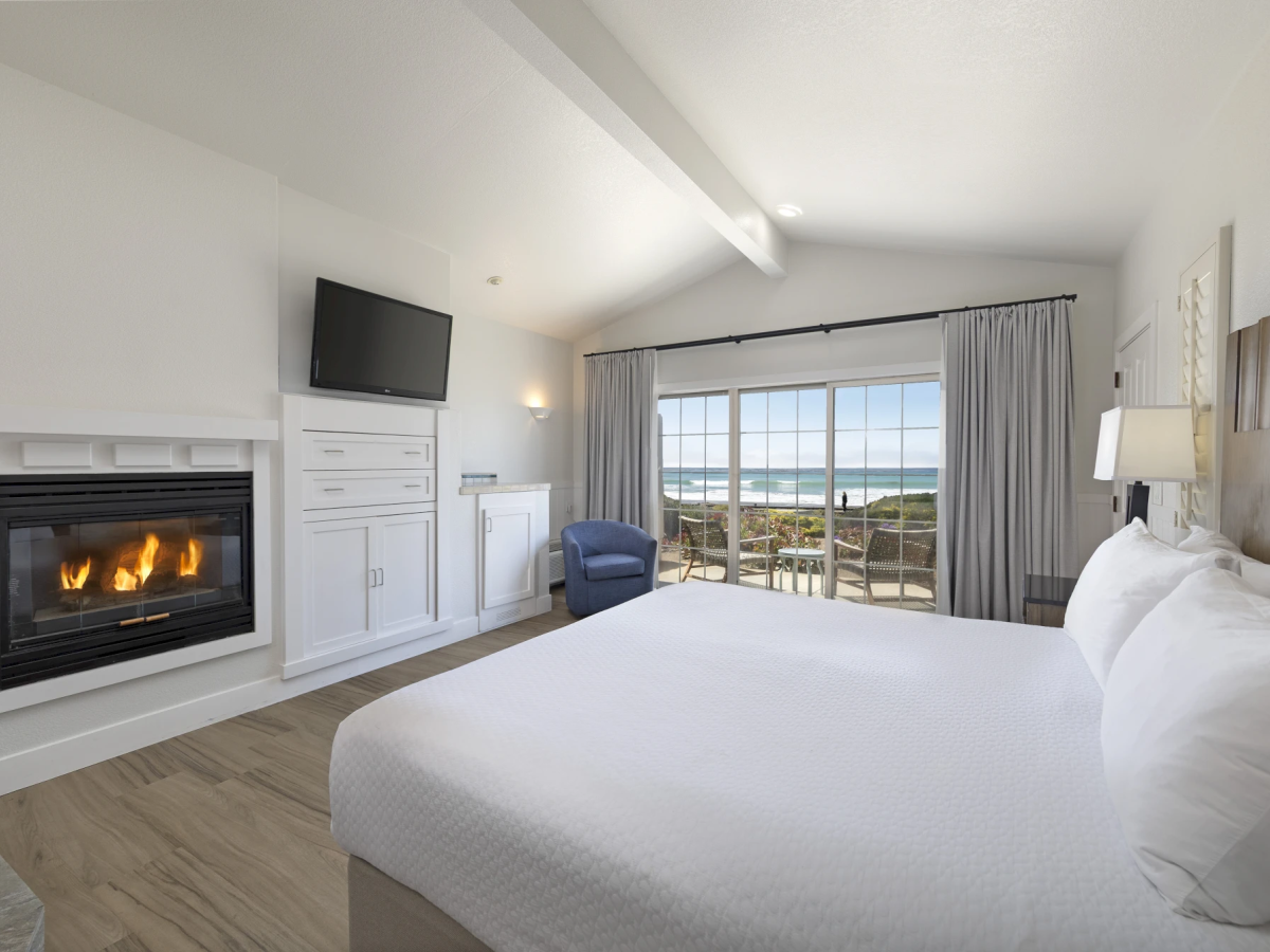 A bright hotel room with a fireplace, built-in white cabinetry, a wall-mounted TV, a comfy blue chair, and a large bed facing glass doors to a balcony with a seascape view.