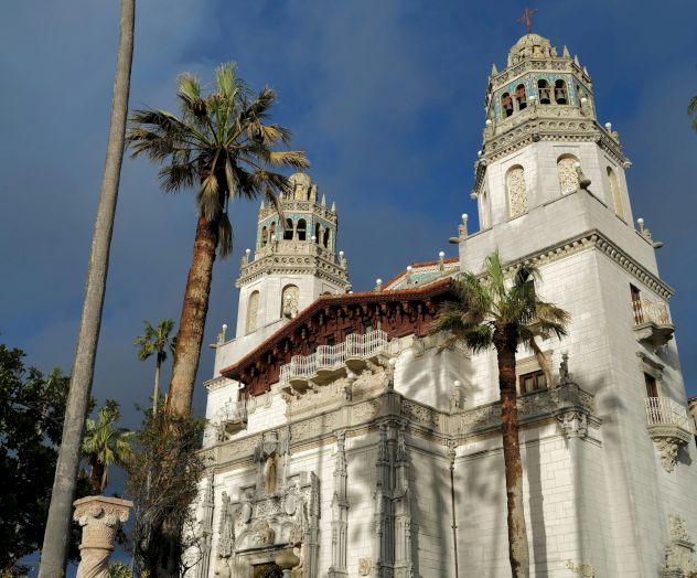 A grand white temple with ornate towers, tall palm trees, and steps leading to an arched entrance against a blue sky.