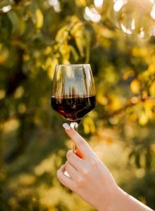 A hand holding a glass of red wine outdoors, with a blurred leafy background. Top it at 140 characters, always ending the sentence.