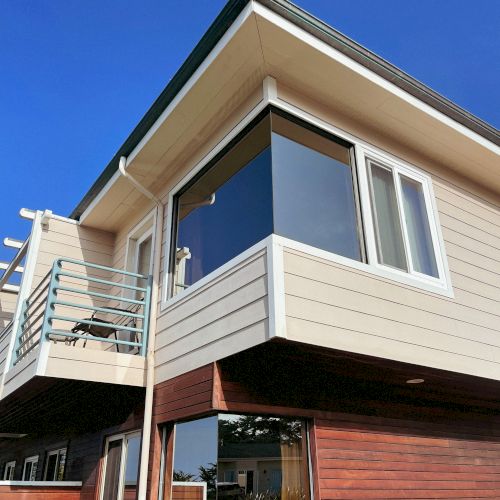 A modern two-story house with large windows, a glass balcony, and mixed wood-and-cladding siding under a bright blue sky.
