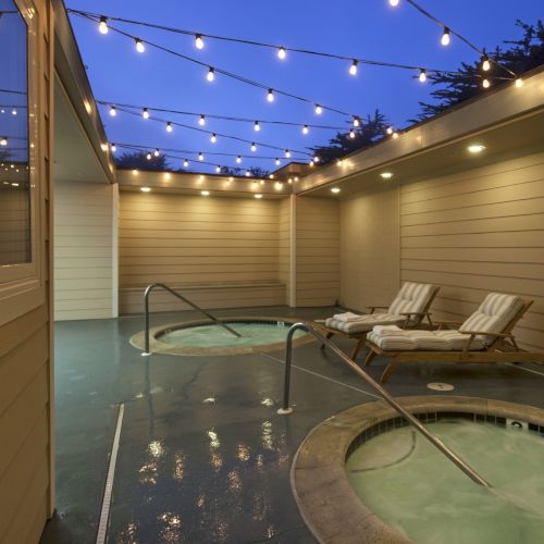 A cozy outdoor spa on a terrace at dusk, with string lights, a small pool, lounge chairs, and warm ambiance.