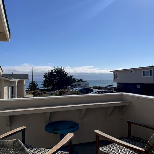 A sunny balcony view with blue sky, sea in the distance, and two wicker chairs facing outward on a small patio.