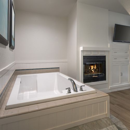 A bright bathroom with a built-in tub, a cozy gas fireplace, and a wall-mounted TV above a white cabinet, all in a clean, modern setting.
