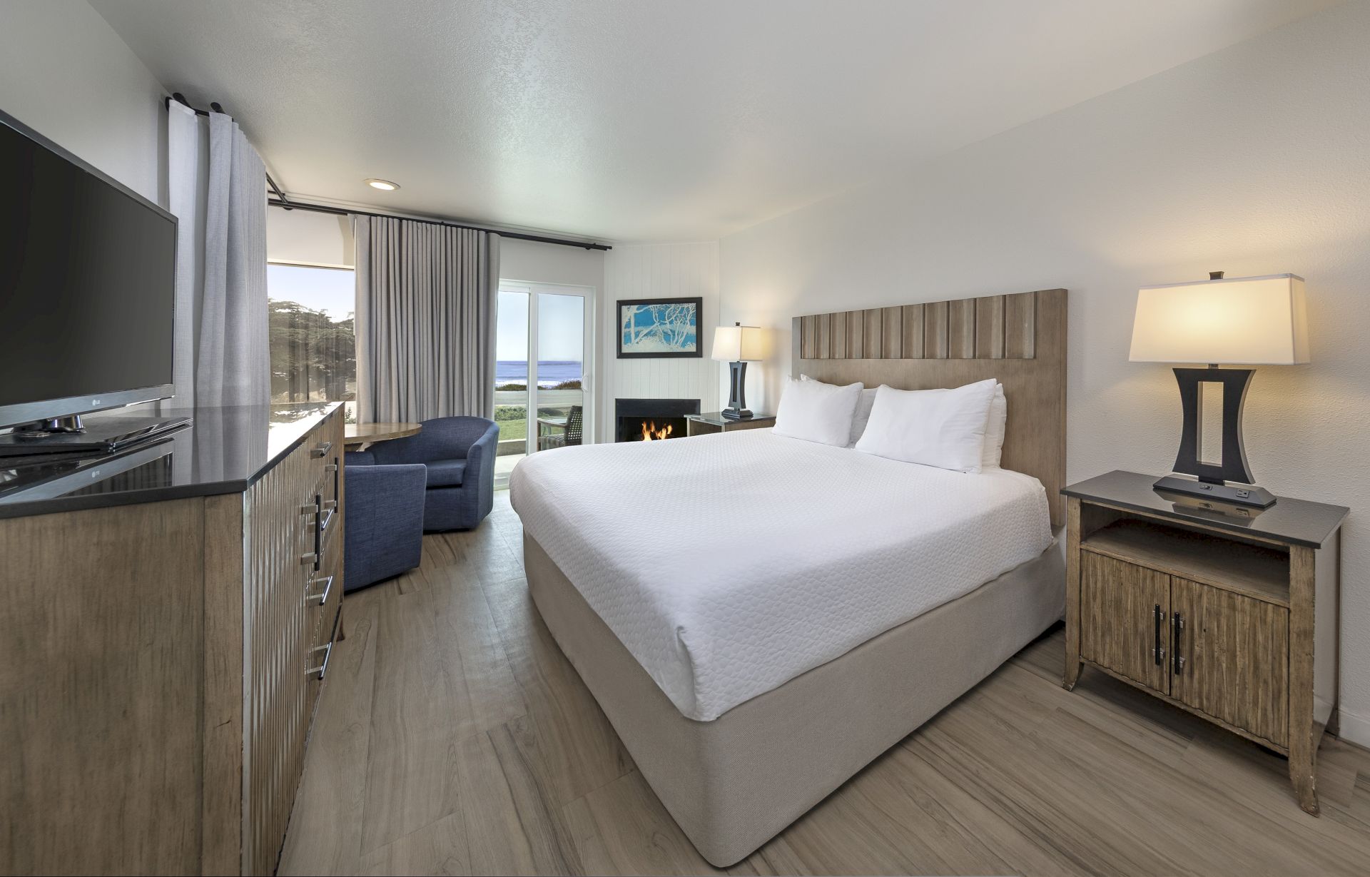 A modern hotel room with a king bed, two nightstands, a dresser with a TV, a seating area, and a sliding glass door to a balcony overlooking the water, bright and airy.