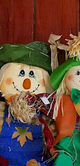 Two handmade scarecrow dolls with straw hair and bright clothes sit beside a large metal jug against a red wooden wall.