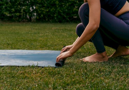 A person is rolling out a yoga mat on grass, crouched near the edge, preparing to exercise outdoors.