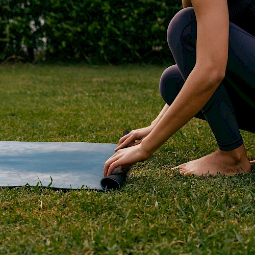 A person is rolling out a yoga mat on grass, crouched near the edge, preparing to exercise outdoors.