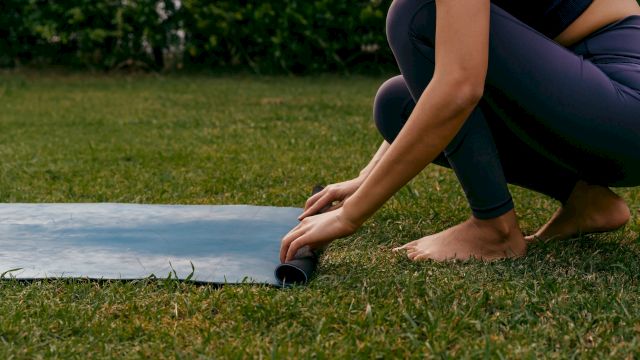 A person is rolling out a yoga mat on grass, crouched near the edge, preparing to exercise outdoors.