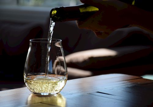 A glass is being filled with light-colored liquor from a bottle; the scene shows a hand pouring into a rounded glass on a wooden table.