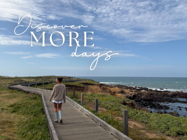 A person strolls along a wooden boardwalk beside rocky cliffs and the ocean under a bright blue sky.