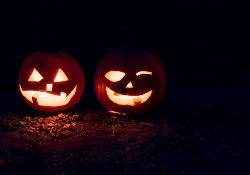 Two glowing Halloween jack-o&rsquo;-lantern pumpkins with carved scary faces sit on a dark ground under a night sky.
