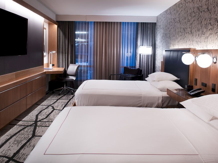 A modern hotel room with two white beds, a desk area, wall-mounted TV, and floor-to-ceiling curtains.