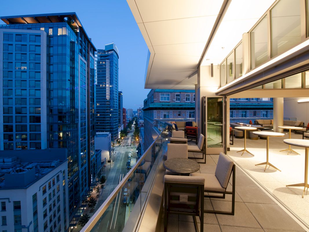 A modern city skyline viewed from a sleek high-rise rooftop lounge with tables and chairs.